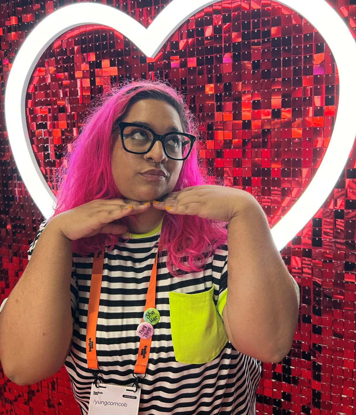 Lissa, with pink hair, puts her hand under chin and looks to the side. Behind her is a large LED heart.